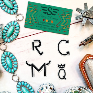 May include: Five black iron-on patches with letters R, C, M, and Q. The Q has a crown on top. The patches are on a white surface with turquoise jewelry, a spur, and a leather holster.