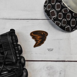 May include: A brown tornado-shaped patch featuring dark brown outlines, positioned between a black leather baseball glove and a black baseball cap with a white football pattern. The logo ES is printed in small black letters below the patch.