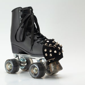 Op de afbeelding: Een zwarte rolschaats met een hoge schoen, zwarte veters en een zilverkleurige metalen plaat. De voorkant van de schaats is voorzien van een zwarte neusbeschermer bedekt met zilverkleurige metalen studs. Het heeft vier zwarte, doorschijnende wielen.