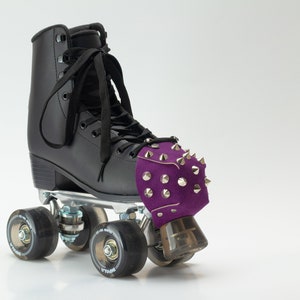 May include: A black roller skate featuring a purple toe guard adorned with silver-toned metal spikes. The skate has black laces, a high-top boot design, and four translucent black wheels attached to a metallic silver base plate.