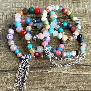 May include: A collection of multi-colored beaded bracelets featuring a variety of round stones in shades of red, blue, green, pink, white, and brown. The bracelets include silver-toned metal chain accents and a dangling silver chain tassel.