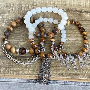 May include: Three bracelets with brown tiger's eye beads and silver chain tassels. The bracelets are layered on a wooden surface. One bracelet is white with a smooth, frosted finish.