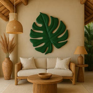 May include: A white sofa with two pillows sits in front of a beige wall with a large green monstera leaf wall decor. A wooden coffee table with a wooden bowl sits in front of the sofa. A wicker lampshade hangs from the ceiling above the sofa. A brown vase with dried pampas grass sits on the floor to the left of the sofa. A brown side table with a brown lamp sits to the right of the sofa.
