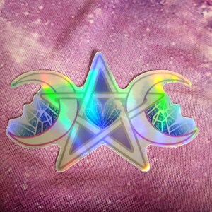 May include: A holographic sticker featuring a triple moon symbol with a pentagram in the center. The sticker is on a purple and white tie-dye fabric background.