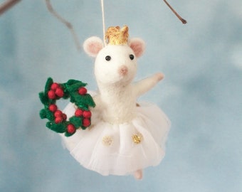 May include: A white felt mouse ornament wearing a gold crown and a white tutu with gold accents. The mouse holds a green wreath with red berries. The ornament hangs from a thin string attached to a brown branch.