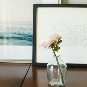 May include: A single pink flower in a clear glass vase on a wooden table. The flower is in focus, and the vase is slightly out of focus. The background is blurred.