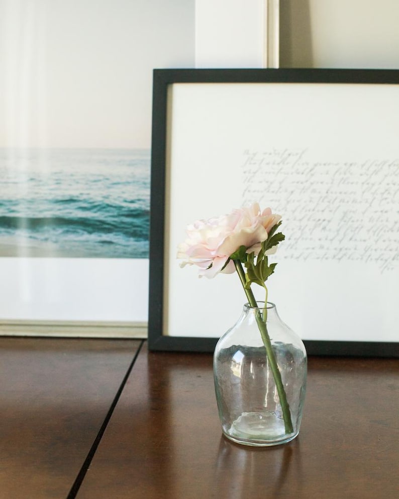 May include: A single pink flower in a clear glass vase on a wooden table. The flower is in focus, and the vase is slightly out of focus. The background is blurred.