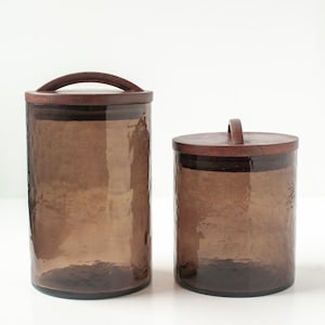 May include: Two cylindrical storage canisters made of translucent brown glass with a hammered texture. Each canister features a circular dark brown wooden lid with a curved handle on top, providing a secure closure for the glass containers.