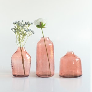 May include: Three pink glass vases of varying sizes and shapes. The vase in the middle is taller and has a flower inside.