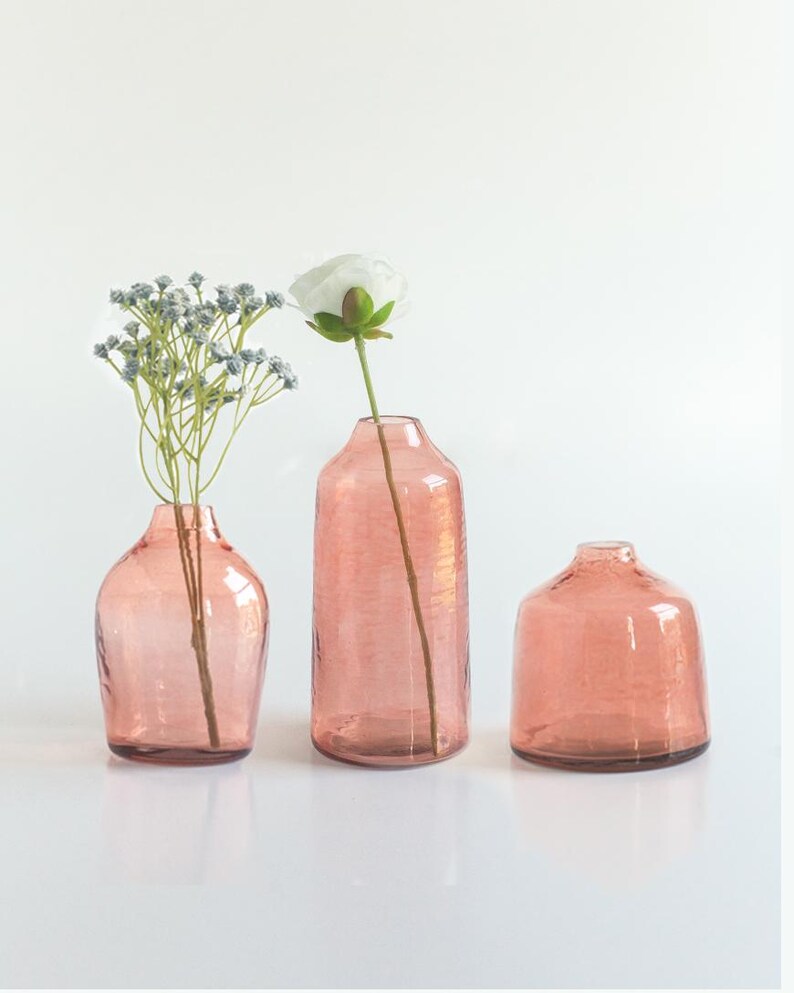 May include: Three pink glass vases of varying sizes and shapes. The vase in the middle is taller and has a flower inside.