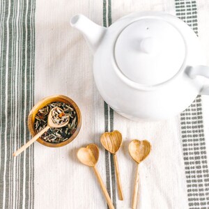May include: A white teapot with a lid and a spout, sitting on a white and gray striped tablecloth. There are three wooden heart-shaped spoons and a small bowl filled with loose leaf tea.