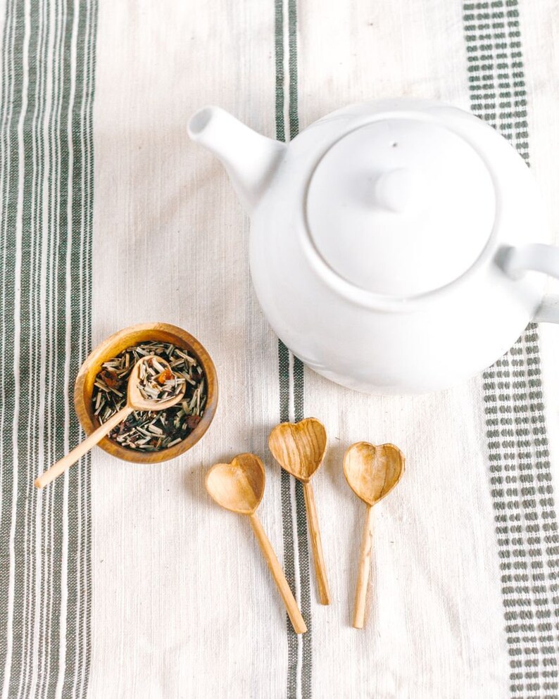 May include: A white teapot with a lid and a spout, sitting on a white and gray striped tablecloth. There are three wooden heart-shaped spoons and a small bowl filled with loose leaf tea.