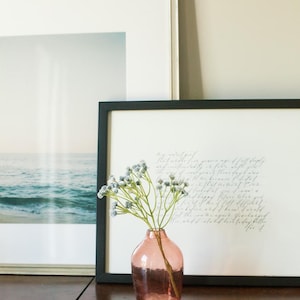 May include: A pink glass vase with a textured surface holds a sprig of dried blue flowers. The vase is sitting in front of two framed prints, one with a seascape and the other with handwritten text.