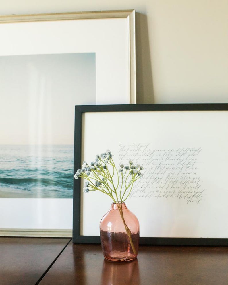May include: A pink glass vase with a textured surface holds a sprig of dried blue flowers. The vase is sitting in front of two framed prints, one with a seascape and the other with handwritten text.