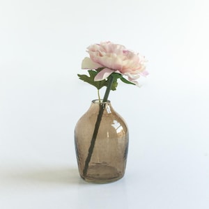 May include: A single pink flower with green leaves in a brown glass vase with a textured surface.