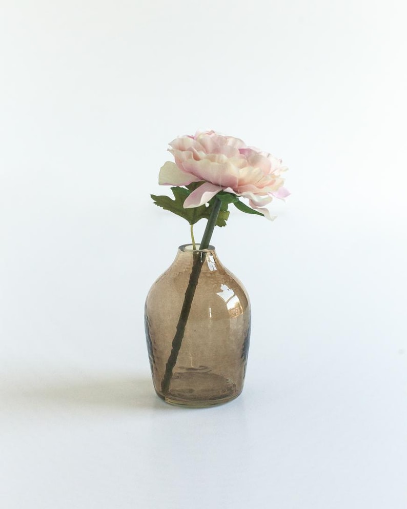 May include: A single pink flower with green leaves in a brown glass vase with a textured surface.
