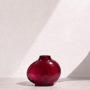 May include: A small, rounded glass vase in a deep, translucent ruby red color. The vessel features a smooth, bulbous body that tapers toward a narrow, circular opening at the top, showcasing a reflective surface with subtle variations in glass thickness.