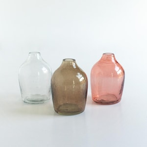 May include: Three glass bud vases in different colours: clear, brown, and pink. The vases are round and have a wide opening at the top.