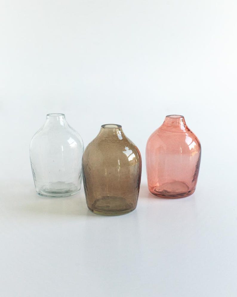 May include: Three glass bud vases in different colours: clear, brown, and pink. The vases are round and have a wide opening at the top.