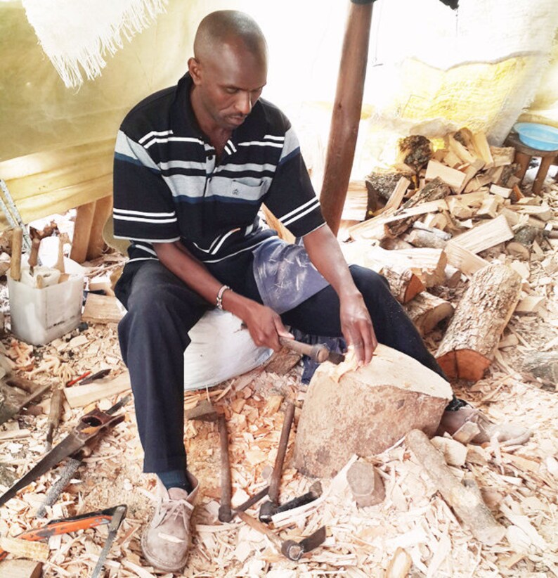 May include: A man is carving wood with a chisel and mallet. He is sitting on a bag of sawdust and surrounded by wood scraps and tools.