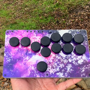 Op de afbeelding: Een aangepaste arcade-vechtstick met een thema van de melkweg. De controller heeft een rechthoekige lay-out met een levendig paars en wit nevelpatroon. Het heeft zwarte knoppen en een joystick om te gamen.