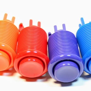 May include: A row of colorful arcade buttons in white, yellow, orange, red, purple, blue, green, and black. Each button has a rounded top and a cylindrical body with a ribbed texture. The buttons are designed for use in arcade games.