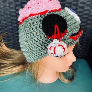 May include: A green crocheted hat with a pink brain, black eye, and white and red eyeball. The hat is on a mannequin head.