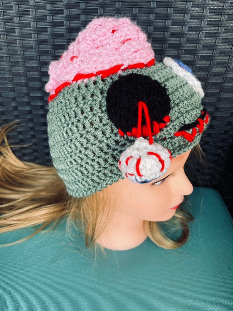 May include: A green crocheted hat with a pink brain, black eye, and white and red eyeball. The hat is on a mannequin head.