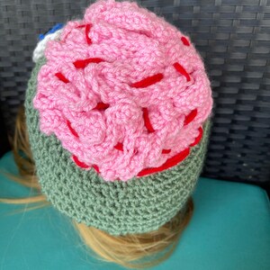 May include: A crocheted hat featuring a pink brain design. The hat is green and has a red outline around the brain.