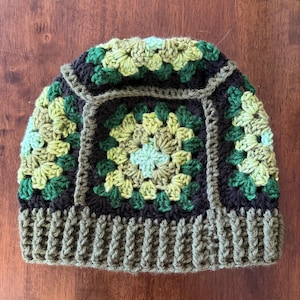May include: A handmade crochet beanie in shades of green and black. The hat features a square granny stitch design in the center and on the sides, with a ribbed brim. The color palette includes olive green, forest green, and black.