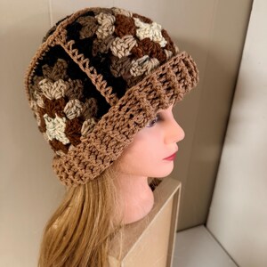 May include: A handmade crochet hat in shades of brown, beige, and black. The hat features a folded brim and a textured, patterned design. The hat is displayed on a mannequin head.