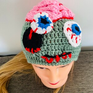 May include: Green crocheted hat with pink, white and red crocheted eyeballs and a black crocheted mouth. The hat is worn on a mannequin with blonde hair.