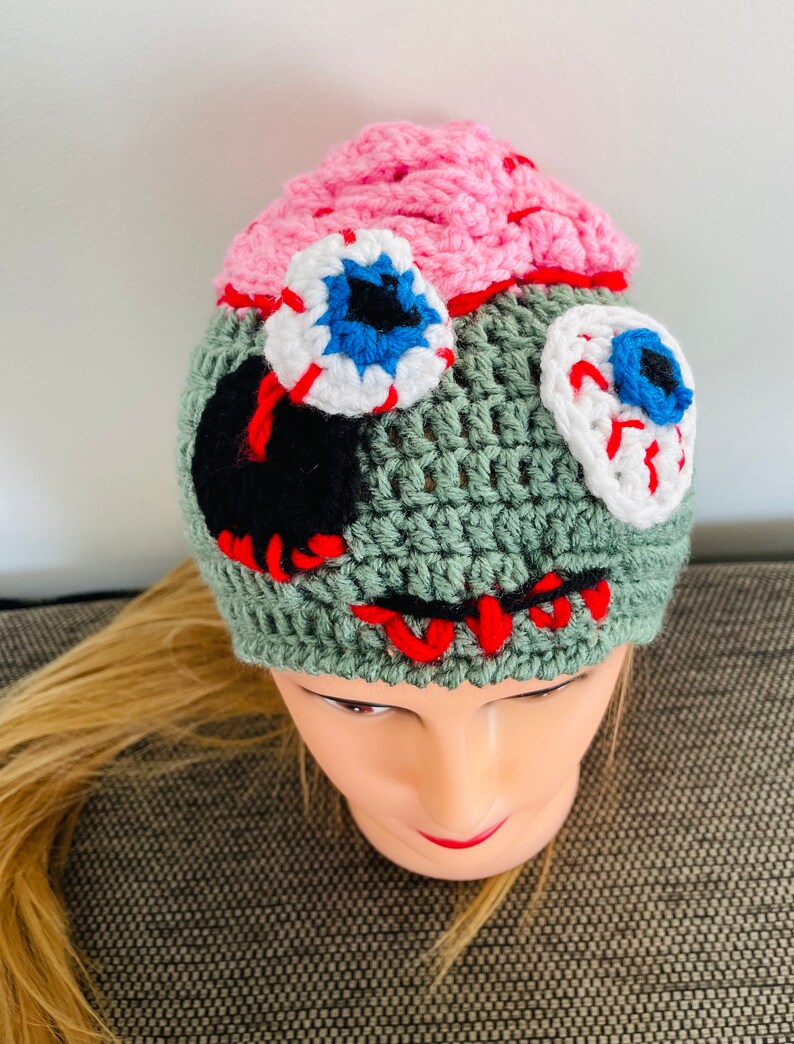 May include: Green crocheted hat with pink, white and red crocheted eyeballs and a black crocheted mouth. The hat is worn on a mannequin with blonde hair.
