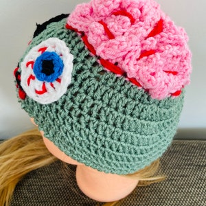 May include: Green crocheted beanie hat with a pink brain, a white eye with a blue iris, and red stitching details.