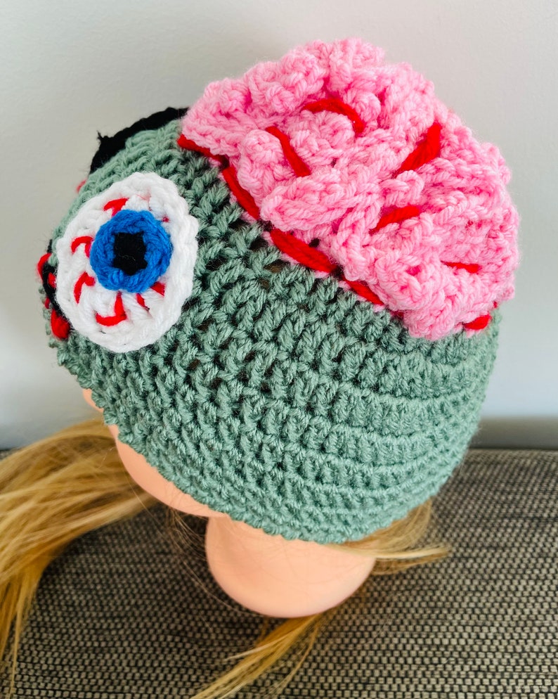 May include: Green crocheted beanie hat with a pink brain, a white eye with a blue iris, and red stitching details.