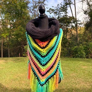 May include: A colorful, handmade crochet shawl with a black cowl neck. The shawl features a vibrant chevron pattern in neon green, yellow, orange, pink, and turquoise, with black stripes. The bottom edge is finished with long, matching fringe.