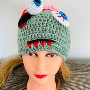 May include: Green crocheted hat with a monster design. The hat has two white eyes with blue and red pupils, a red mouth, and black stitching.