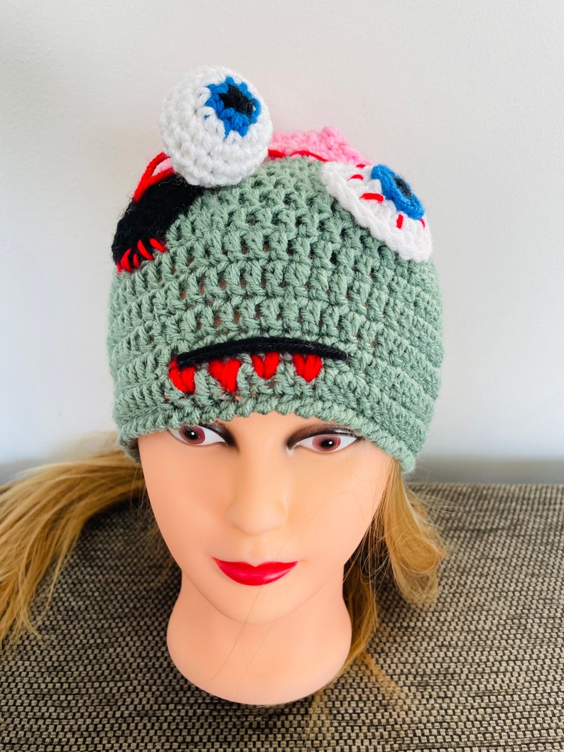 May include: Green crocheted hat with a monster design. The hat has two white eyes with blue and red pupils, a red mouth, and black stitching.