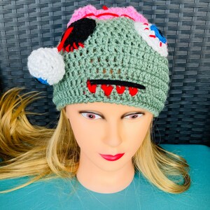 May include: Green crocheted hat with a stitched-on face, featuring a pink brain, black eye, white eye with a blue pupil, and red teeth.