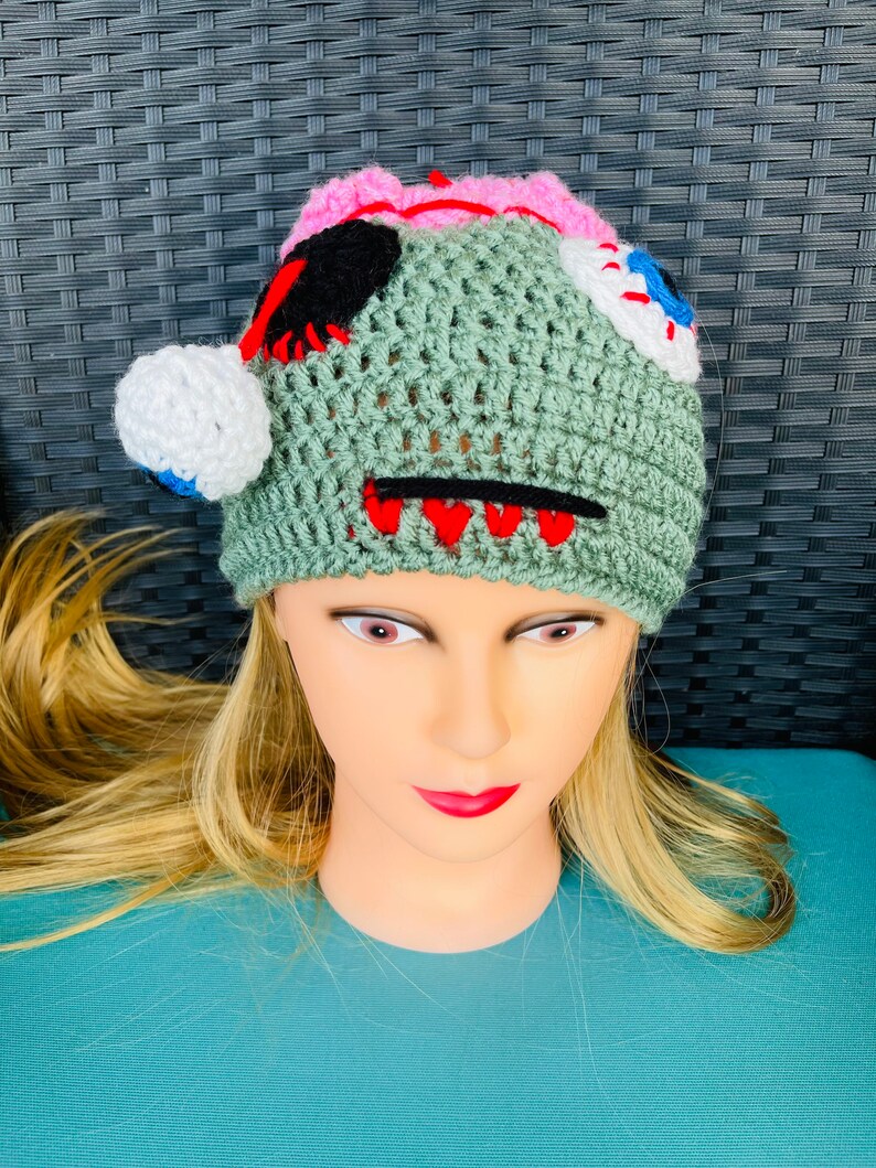 May include: Green crocheted hat with a stitched-on face, featuring a pink brain, black eye, white eye with a blue pupil, and red teeth.