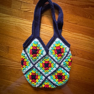 May include: A crocheted tote bag with a navy blue strap and a colorful geometric pattern. The bag is made of yarn in shades of red, yellow, green, and white.