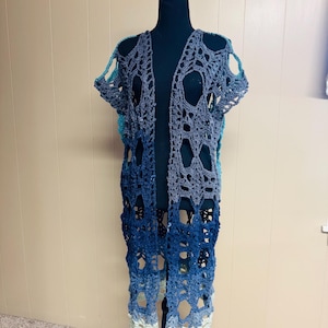May include: A long, open-front crochet cardigan in shades of blue and gray, displayed on a black mannequin. The cardigan features an open, lacy design with a gradient effect, transitioning from light blue at the bottom to darker shades at the top. The sleeves are short.