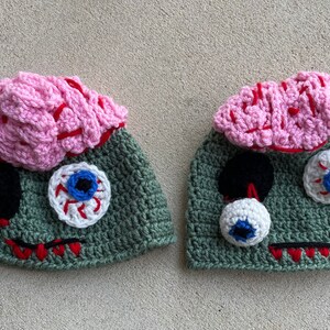 May include: Two green crocheted hats with pink brains and googly eyes. The hats are designed to look like zombies.