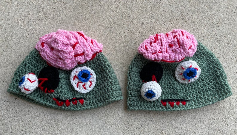 May include: Two green crocheted hats with pink brains and googly eyes. The hats are designed to look like zombies.