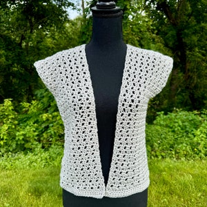 May include: A light gray, open-front crochet vest with short sleeves. The vest is displayed on a black mannequin against a backdrop of green foliage. The crochet pattern features an open, airy design.