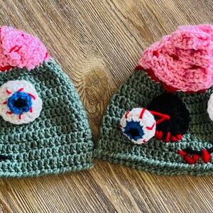 May include: Two green crocheted hats with pink brains and googly eyes. The hats have red stitching and black details.