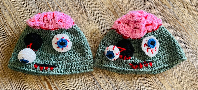 May include: Two green crocheted hats with pink brains and googly eyes. The hats have red stitching and black details.
