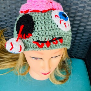 May include: Green crocheted hat with pink, white and black details. The hat has a stitched-on eye with a blue iris and white sclera. The hat is designed to look like a zombie.