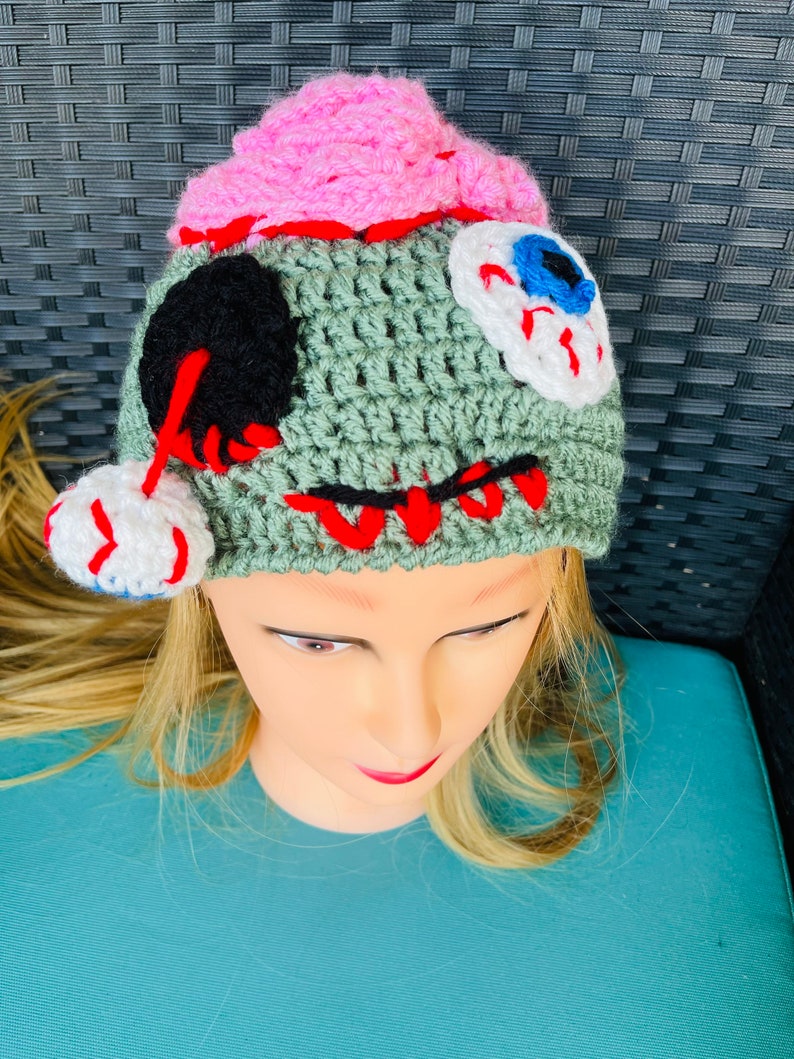 May include: Green crocheted hat with pink, white and black details. The hat has a stitched-on eye with a blue iris and white sclera. The hat is designed to look like a zombie.