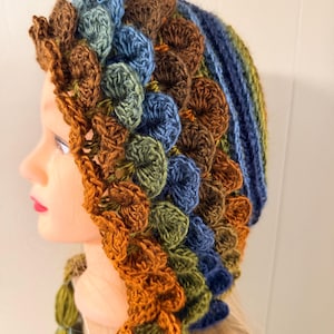 May include: A handmade crochet hat in shades of brown, blue, and green. The hat features a textured, scale-like design around the crown and a decorative trim. The hat has a tassel detail on the side.